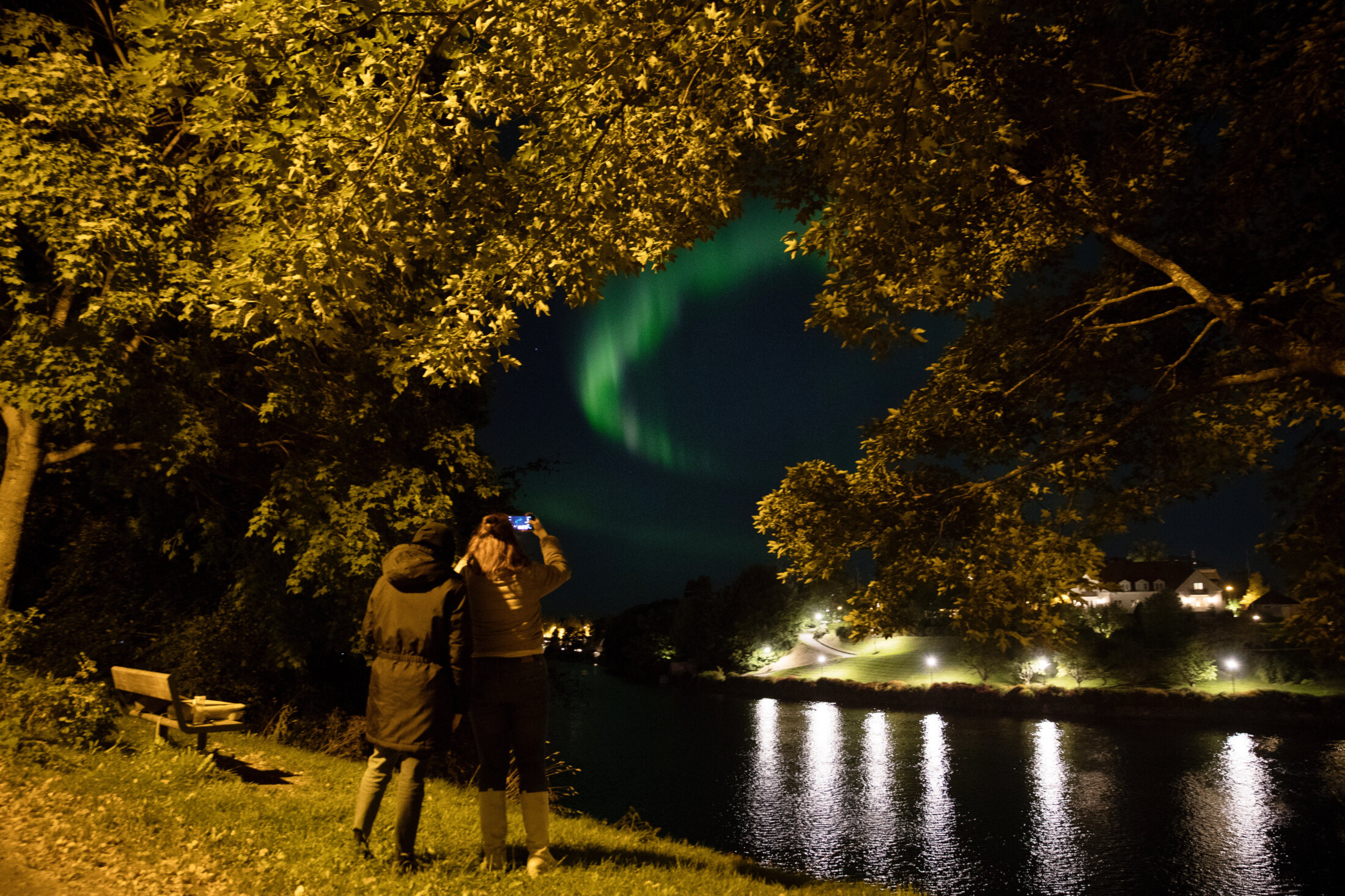 To personer står ved Nidelva og fotograferer nordlyset mellom trærne